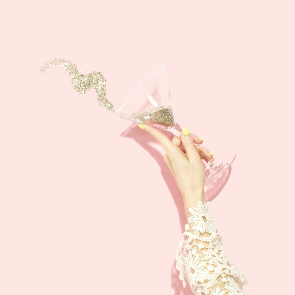 A Hand Tossing Glitter Out of a Cocktail Glass Against a Pink Background