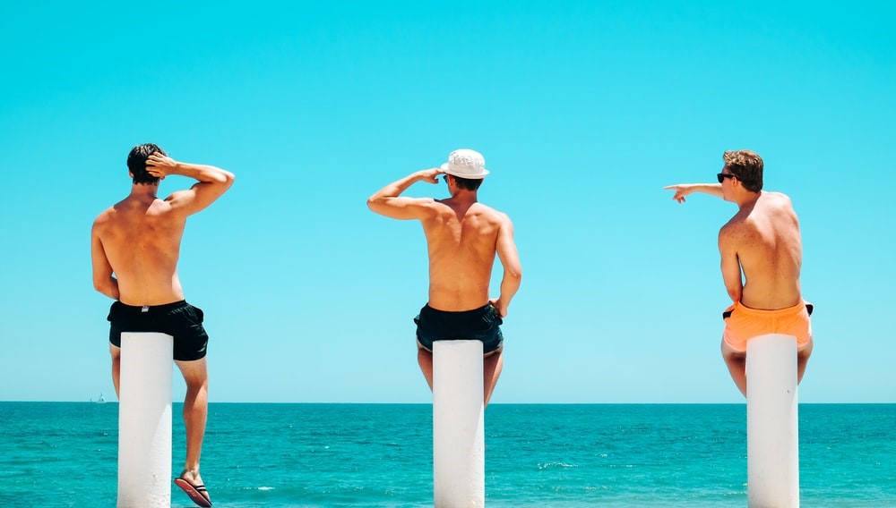 Three Shirtless Men Sitting on Concrete Poles by the Beach
